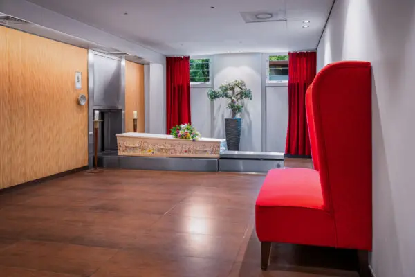 Crematieruimte met houten wandafwerking, een doorgang en een rode stoel op de voorgrond.