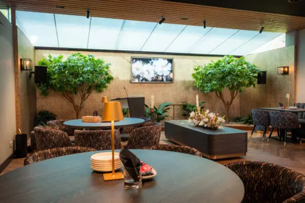 Koffiekamer met ronde tafels, planten en een groene wand met beeldscherm.