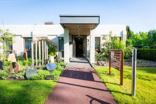 Hoofdingang van Crematorium Kranenburg met glazen overkapping en groen rondom.