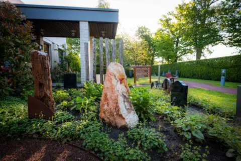 Houten gedenkobject en stenen gedenkobject in het groen naast Crematorium Kranenburg.