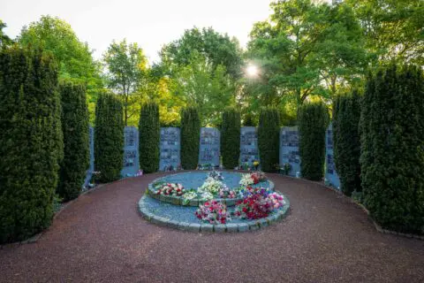 Ronde gedenkplaats met bloemen, omringd door gedenkpanelen en hoge groene hagen.